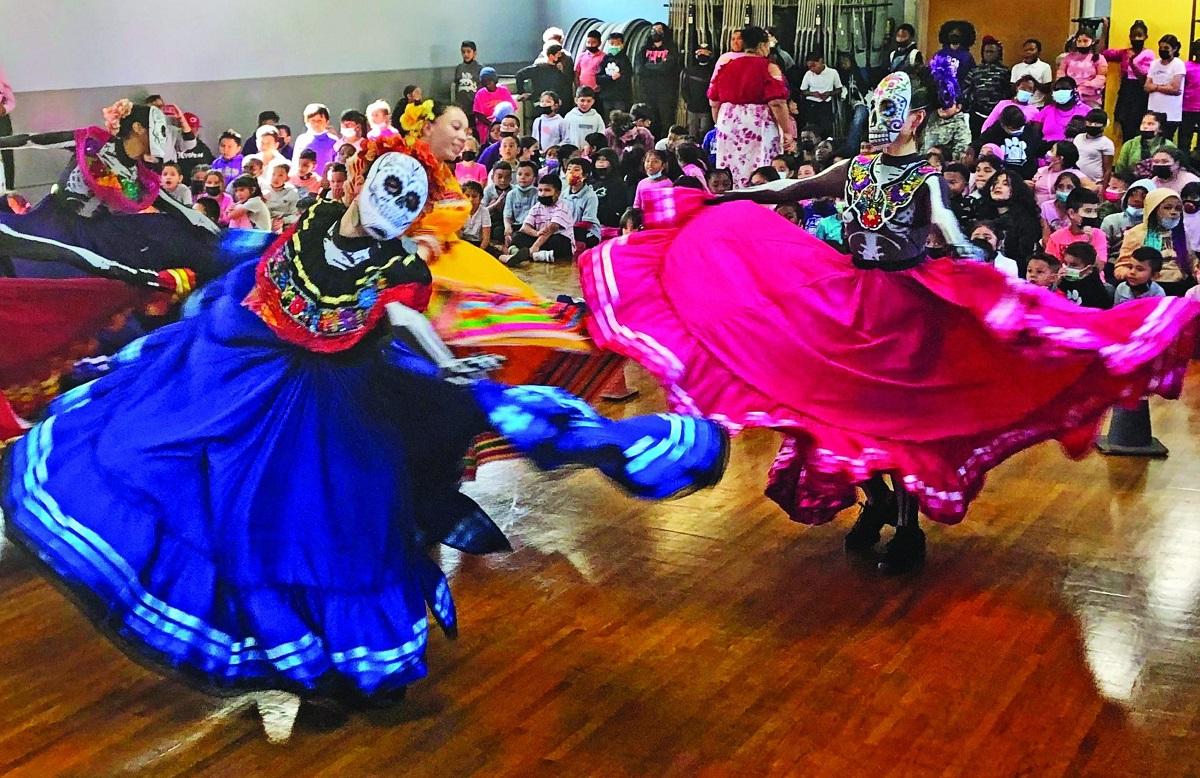 Female dancers, wearing masks decorated as skulls, turn about the room in colorful, full skirts as students watch.