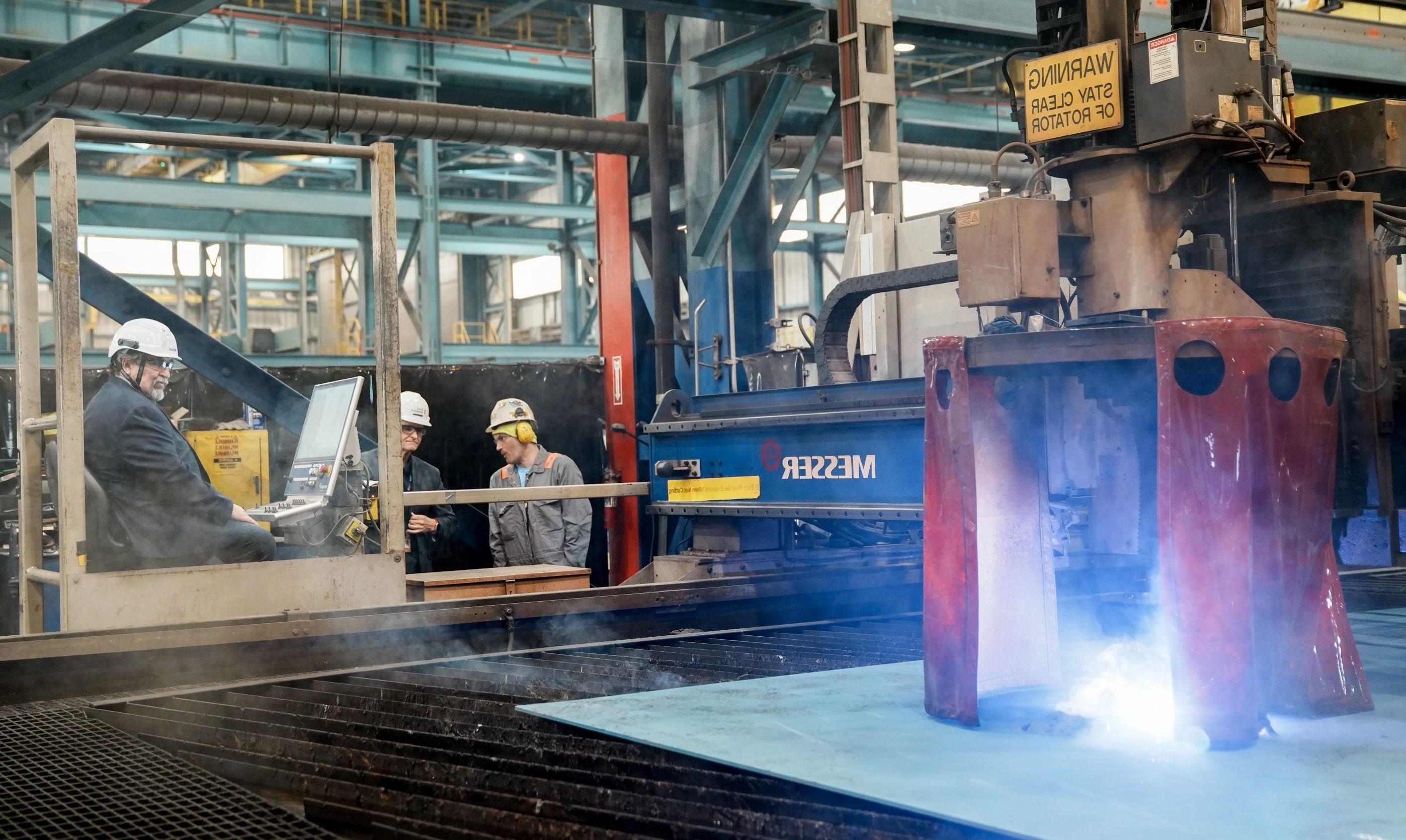 A bright light comes from the cutting machine as the first piece of steel is cut and a 澳博体育app下载 employee wearing a hard hat sits behind the controls. 