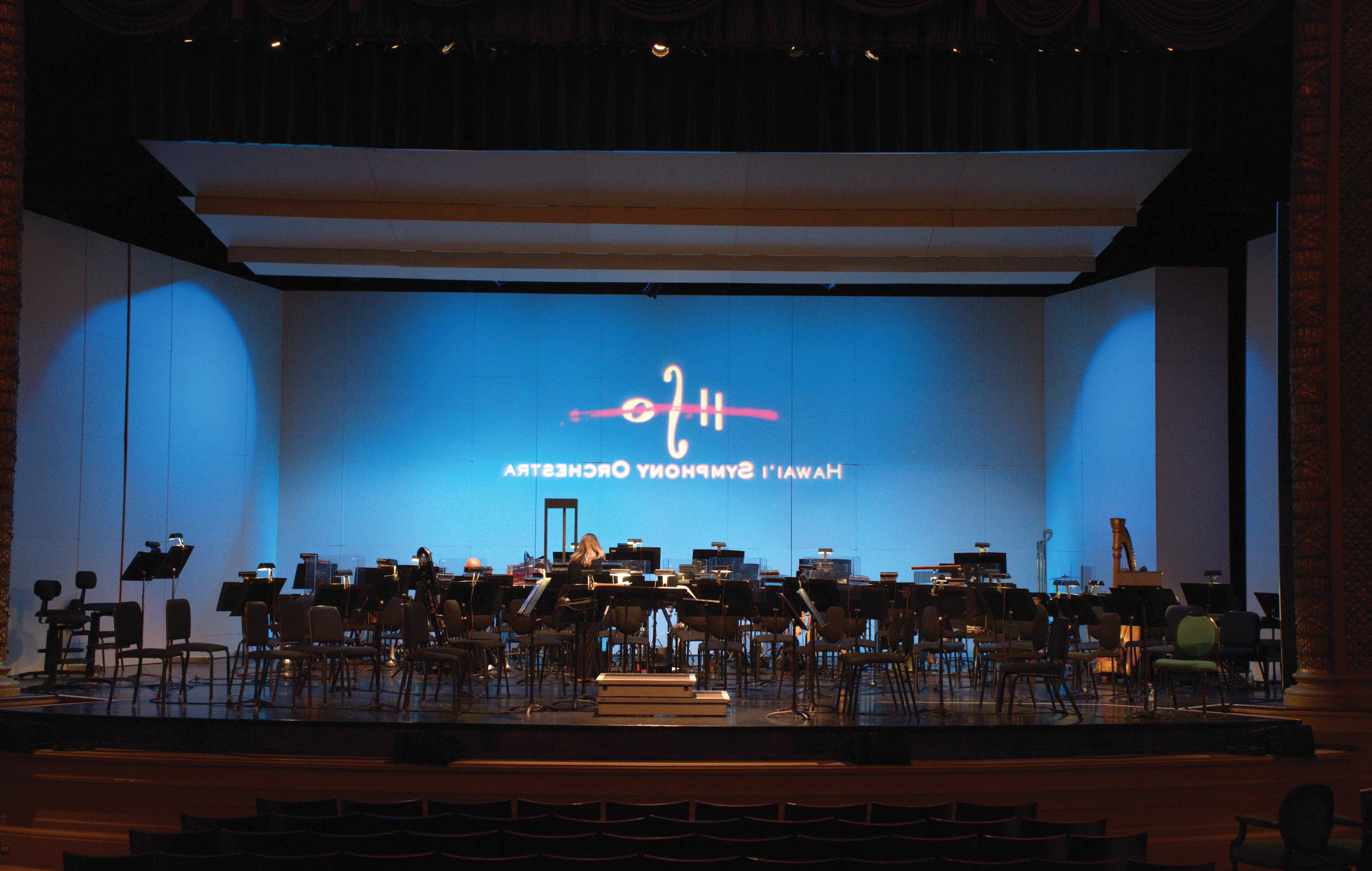 The Hawaii Symphony Orchestra logo shines on the back on the stage which is set up with chairs and music stands for the symphony. The light colored accoustic shell hangs from the ceiling and surrounds the sides and rear of the stage.